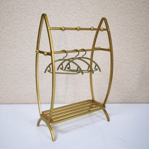 May include: A gold-colored metal clothing rack featuring an arched frame, two horizontal bars with small spherical accents, a slatted base, and three matching gold-colored metal hangers suspended from the middle horizontal bar.