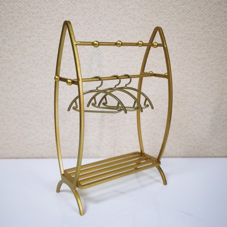 May include: A gold-colored metal clothing rack featuring an arched frame, two horizontal bars with small spherical accents, a slatted base, and three matching gold-colored metal hangers suspended from the middle horizontal bar.