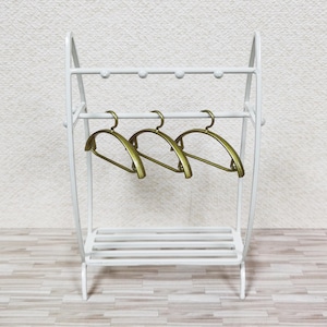 May include: A white metal clothing rack featuring a top horizontal bar with four small rounded knobs and a lower slatted shelf. Three metallic gold-toned hangers are suspended from the middle horizontal bar of the freestanding structure.
