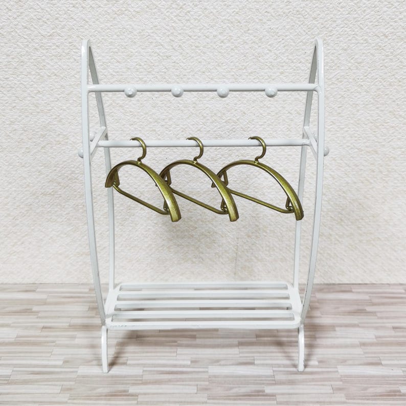 May include: A white metal clothing rack featuring a top horizontal bar with four small rounded knobs and a lower slatted shelf. Three metallic gold-toned hangers are suspended from the middle horizontal bar of the freestanding structure.