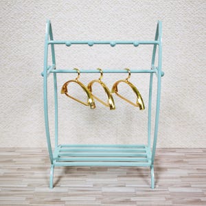 May include: A light blue metal clothing rack with a curved frame, a top bar featuring five small rounded knobs, and a slatted bottom shelf. Three shiny gold-toned hangers are suspended from the middle horizontal bar of the rack.