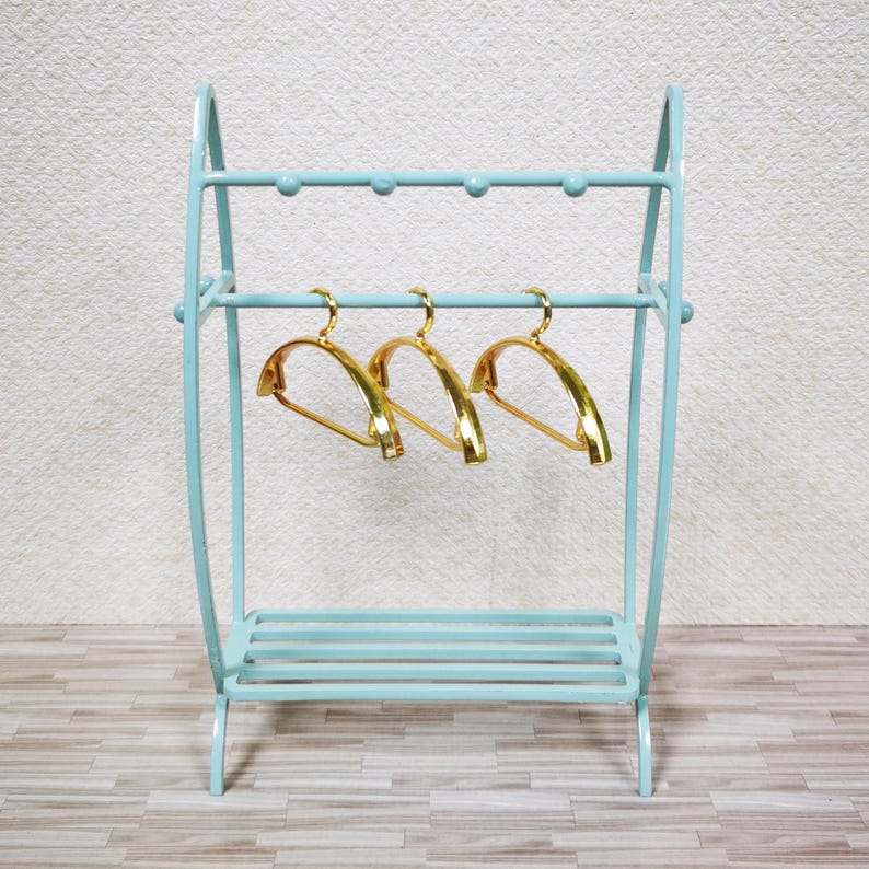 May include: A light blue metal clothing rack with a curved frame, a top bar featuring five small rounded knobs, and a slatted bottom shelf. Three shiny gold-toned hangers are suspended from the middle horizontal bar of the rack.