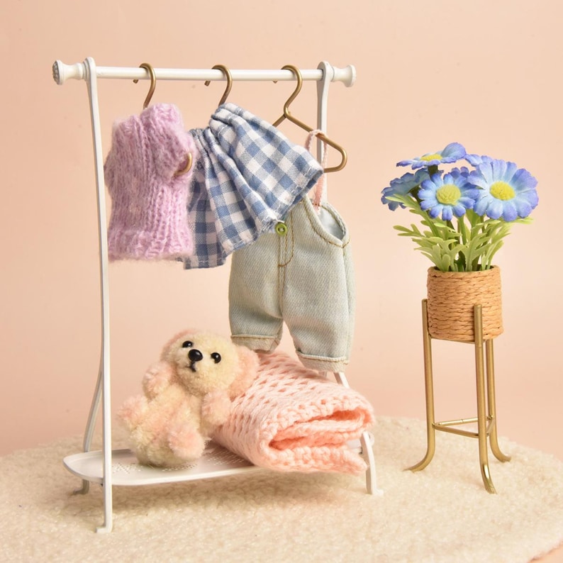 May include: A white metal clothing rack holding a miniature lavender knit sweater, a blue and white gingham skirt, and light blue denim overalls on gold hangers. A small plush bear and a folded pink knit blanket sit on the base shelf.
