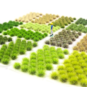 Peut inclure: Une scène miniature de paysage avec des touffes d'herbe modèle vertes et beiges. Une petite figurine d'une personne avec un chapeau jaune et une chemise bleue se tient dans la scène, suggérant un modèle réduit ou un diorama.