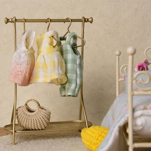 Può includere: Un appendiabiti in metallo dorato con tre grucce che tengono un vestito a quadri rosa, giallo e verde. Una piccola borsa di vimini si trova sullo scaffale inferiore dell'appendiabiti. Un letto bianco con una trapunta a motivi floreali si trova sullo sfondo.