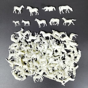 Puede incluir: Una colección de pequeñas figuras de caballos de plástico blanquecino en varias poses, incluyendo de pie, pastando y corriendo. Las figuras están dispersas sobre una superficie gris oscuro, creando una escena juguetona y caprichosa.