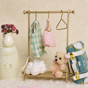 May include: A gold-toned metal clothing rack holding a green and white checkered jumpsuit and a pink crocheted dress on hangers. A small beige teddy bear sits on the base shelf next to a white folded fabric item and a teal floral suitcase.