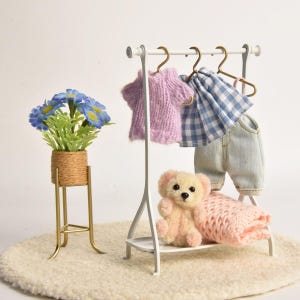 May include: A white metal clothing rack holding a miniature purple knit sweater, a blue and white gingham skirt, and light blue denim pants. A small beige teddy bear sits on the rack base next to a folded pink knit blanket and a blue flower pot.