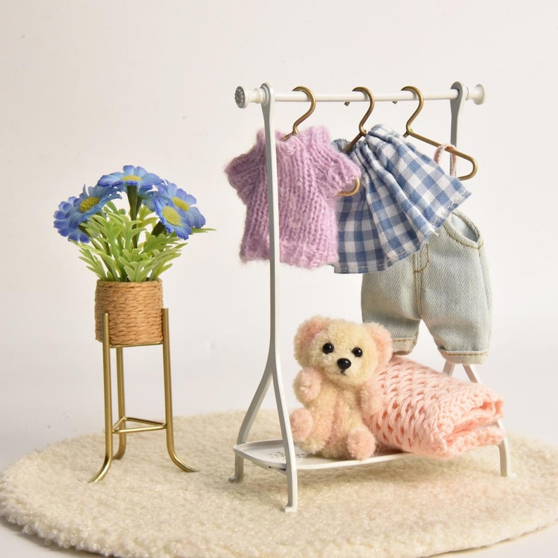 May include: A white metal clothing rack holding a miniature purple knit sweater, a blue and white gingham skirt, and light blue denim pants. A small beige teddy bear sits on the rack base next to a folded pink knit blanket and a blue flower pot.