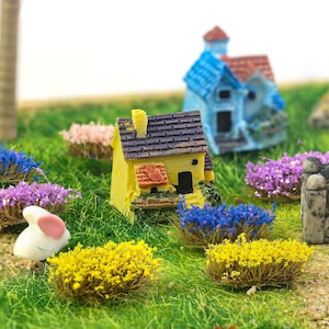 Peut inclure: Une scène de jardin de fées miniature avec une maison jaune et un toit gris foncé, un lapin blanc et des buissons miniatures colorés. La scène est sur de l'herbe verte, avec une maison bleue en arrière-plan.