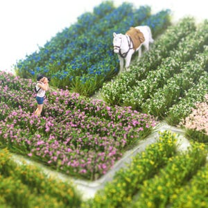 May include: Miniature diorama of a field with rows of colorful flowers and grass. A small figure of a person stands in the pink flowers, and a white horse with a saddle is in the background. The scene is set against a white backdrop.