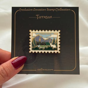 Può includere: Spilla rettangolare in smalto a forma di francobollo. La spilla presenta un paesaggio montano con un castello, alberi e un cervo. Il testo "Terrasen" e "600" sono visibili. La spilla è su una carta nera con la scritta "Exclusive Location Stamp Collection".