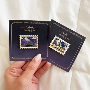 Puede incluir: Dos pines de esmalte dorado con una ilustración estilizada de un horizonte urbano con montañas y una luna creciente. Los pines están en tarjetas con el texto "Exclusive Location Stamp Collection: Velaris the city of stars" y las direcciones de los sitios web "sallteas.co" y "iteas.com".