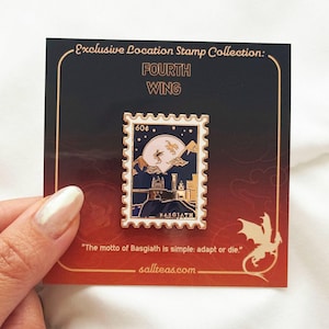 Op de afbeelding: Een gouden emaille pin met een postzegelontwerp op een blauwe achtergrond. De postzegel toont een kasteel met een draak die eroverheen vliegt, een volle maan en sterren. De tekst "BASGIATH" en "60¢" staat op de postzegel gedrukt. De pin wordt getoond op een kaart met de tekst "Exclusive Location Stamp Collection: FOURTH WING" en "Het motto van Basgiath is simpel: aanpassen of sterven."