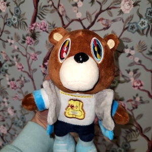 Puede incluir: Un oso de peluche marrón con ojos coloridos, que lleva una camisa blanca con un gráfico amarillo, una chaqueta gris, pantalones azules y zapatos azules. El oso tiene una nariz negra y un hocico blanco. Un artículo de colección.
