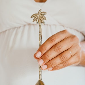 May include: A silver palm tree spoon with a rounded bowl. The spoon is held in a hand.