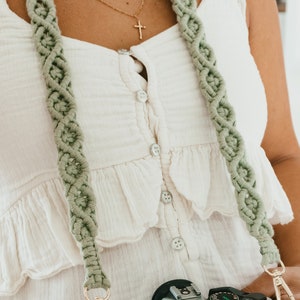 Puede incluir: Una correa de cámara verde hecha de macramé con un cierre de metal dorado unido a una cámara plateada. La cámara tiene la palabra "Canon" impresa en ella.