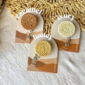 Puede incluir: Tres carretes de insignia redondos de ratán tejido en colores caramelo, marfil y miel. Cada carrete está unido a un porta-insignia de plástico transparente y un cordón retráctil plateado. Los carretes se muestran en tarjetas individuales con el nombre de la marca "SIENNAF*X" impreso en ellas.