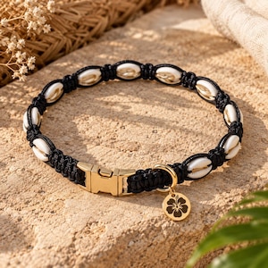 May include: A black macrame collar featuring white cowrie shells woven into the cord. The collar includes a gold-toned metal side-release buckle and a circular gold-toned metal charm engraved with a hibiscus flower graphic.