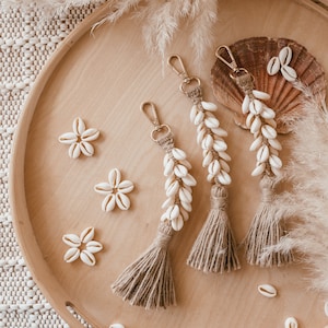 Può includere: Tre portachiavi dorati con nappe marroni e conchiglie bianche infilate sulla catena. I portachiavi sono esposti su un vassoio di legno con conchiglie bianche sparse intorno.