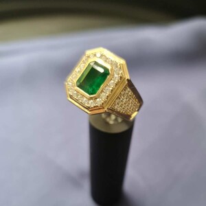 Op de afbeelding: Een gouden ring met een grote smaragdgroene edelsteen omringd door kleinere heldere edelstenen. De ring staat op een zwarte standaard.