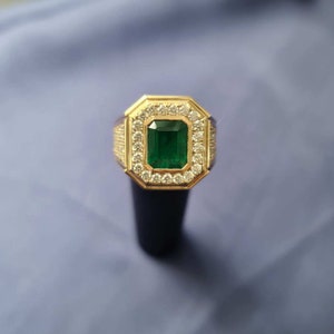 Op de afbeelding: Een gouden ring met een grote smaragdgroene edelsteen omringd door kleinere heldere edelstenen.