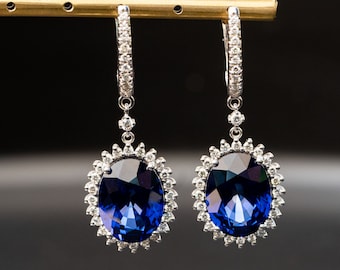 May include: A pair of silver earrings with a large oval sapphire gemstone surrounded by smaller diamonds. The earrings are hanging from a silver chain.