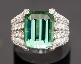 May include: A ring with a large emerald-cut green gemstone surrounded by a band of smaller diamonds.