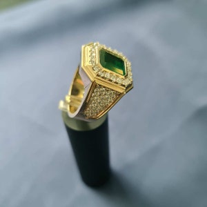 Op de afbeelding: Een gouden ring met een grote smaragdgroene edelsteen omringd door kleinere heldere edelstenen. De ring staat op een zwarte standaard.