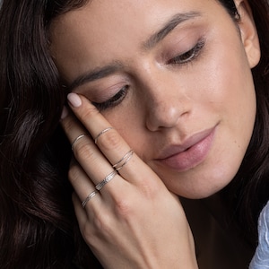 Könnte beinhalten: Eine Frauenhand trägt mehrere dünne Silberringe. Die Ringe sind an jedem Finger übereinander gelegt und haben ein zartes, strukturiertes Design.