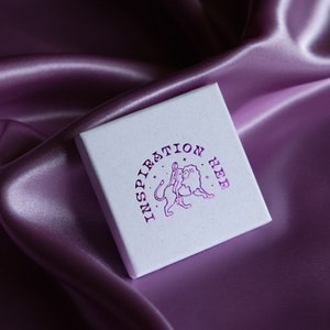 May include: A white box with the words "Inspiration Her" printed in pink on the top. The box has a graphic of a woman standing next to a lion.