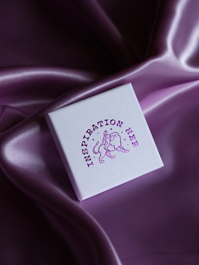 May include: A white box with the words "Inspiration Her" printed in pink on the top. The box has a graphic of a woman standing next to a lion.