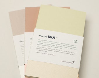 May include: A stack of three notebooks with different colored covers. The top notebook is light green, the middle notebook is light pink, and the bottom notebook is light beige. The notebooks are made of recycled paper and have the text "Hey, I'm NOLA" printed on the front. The text is in black and white. The notebooks are sitting on a light gray surface.