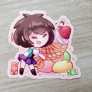 Puede incluir: Una pegatina con un personaje de dibujos animados con cabello casta&ntilde;o, que lleva una camisa blanca y una falda de cuadros morados, sosteniendo una criatura verde y azul. El personaje est&aacute; de pie junto a un gran postre colorido con una fresa en la parte superior. El postre est&aacute; rodeado de chispas y otros peque&ntilde;os caramelos.