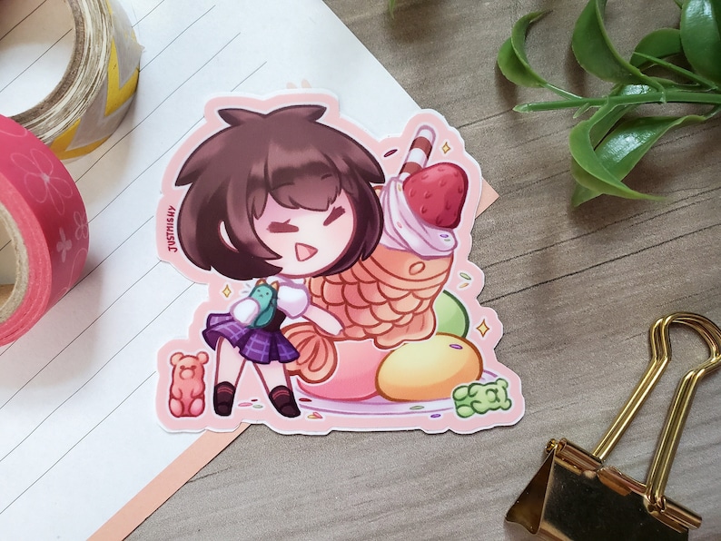 Puede incluir: Una pegatina con una ni&ntilde;a de dibujos animados de pelo casta&ntilde;o sosteniendo un gran taiyaki relleno de helado y una fresa. El taiyaki est&aacute; rodeado de caramelos y chispas. La pegatina tiene un fondo rosa y el texto "JustSushi" es visible en la esquina inferior izquierda.