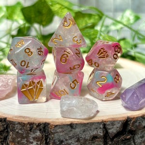 May include: A set of polyhedral dice featuring a translucent, milky base with swirls of pink and blue, accented by gold-coloured numbers and symbols. The set includes various shapes, including a d20, d12, d10, d8, d6, and d4, alongside two small, smooth, unnumbered crystal-like stones.