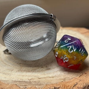 以下が含まれることがあります： ステンレス製のティーインフューザーと、虹色の多面体サイコロ。サイコロには白い数字が書かれています。ティーインフューザーは、ヒンジ付きの留め金が付いたメッシュボールです。どちらのアイテムも木製の表面に置かれています。