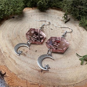May include: Earrings with pink, faceted dice charms and silver hooks. Each earring has a silver crescent moon and star charm. The earrings are displayed on a wooden surface. The dice are numbered.