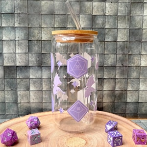 Könnte beinhalten: Ein durchsichtiger Glasbecher mit Bambusdeckel und Strohhalm. Das Glas ist mit lila geometrischen Formen, Fledermäusen und Sternen verziert. Mehrere lila Würfel sind auf einer Holzscheibe vor dem Glas verstreut. Der Becher ist für kalte Getränke.