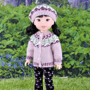 Op de afbeelding: Een pop die een roze en groene gebreide trui draagt met een witte knoopsluiting, een roze en bruine gebreide muts en zwarte leggings met een wit bloemenpatroon. De pop staat voor een groene, bladerrijke achtergrond.