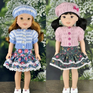 Op de afbeelding: Twee poppen die gebreide vesten en bloemenrokken dragen. De pop links draagt een blauw-wit gestreept vest en een blauwe bloemenrok met witte kant. De pop rechts draagt een roze vest en een groene bloemenrok met witte kant.