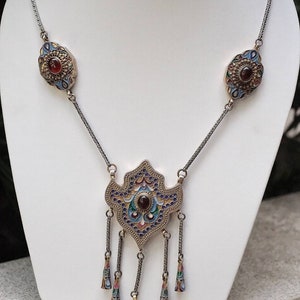 Puede incluir: Un collar de plata con un gran colgante adornado con un dise&ntilde;o de esmalte azul, dorado y rojo. El colgante tiene una piedra preciosa roja en el centro y cuelga de una cadena con colgantes m&aacute;s peque&ntilde;os a juego.
