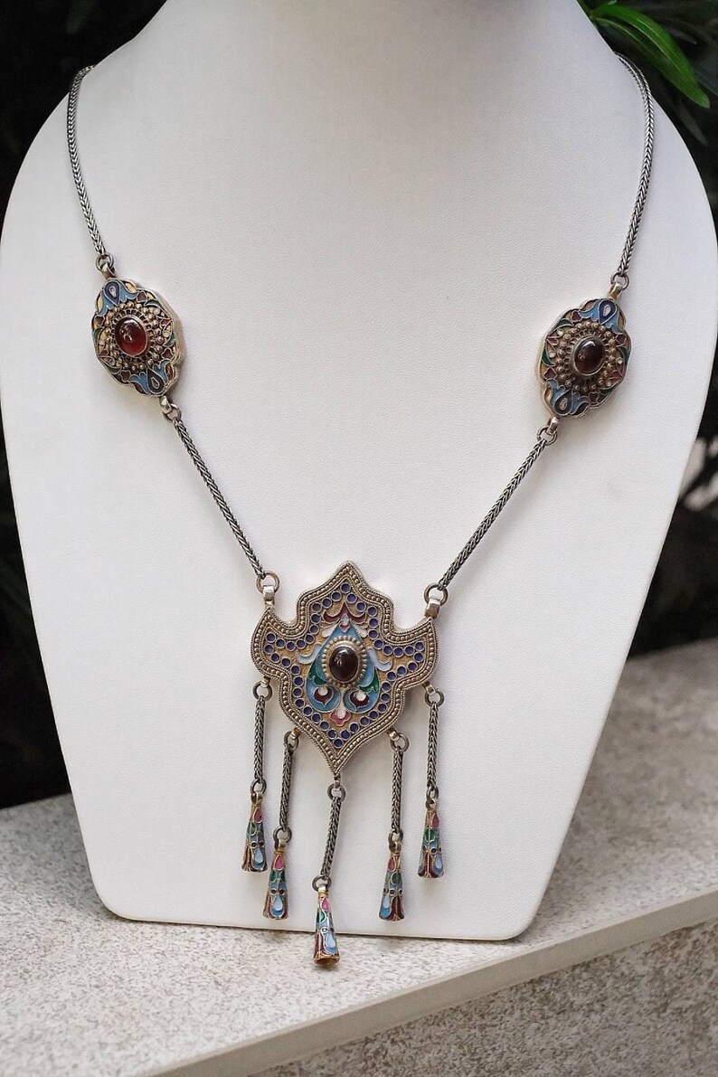 Puede incluir: Un collar de plata con un gran colgante adornado con un dise&ntilde;o de esmalte azul, dorado y rojo. El colgante tiene una piedra preciosa roja en el centro y cuelga de una cadena con colgantes m&aacute;s peque&ntilde;os a juego.