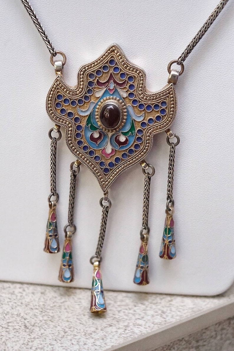 Puede incluir: Un collar de plata con un gran colgante adornado. El colgante est&aacute; hecho de plata y esmalte y presenta un dise&ntilde;o geom&eacute;trico con acentos azules, verdes, rojos y dorados. El colgante est&aacute; unido a una cadena de plata con cinco colgantes m&aacute;s peque&ntilde;os que cuelgan.