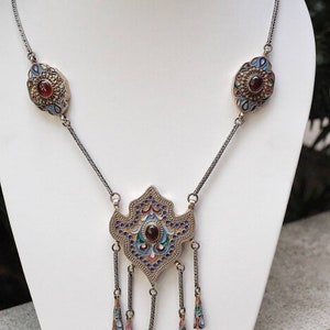 Puede incluir: Un collar de plata con un gran colgante adornado con trabajo de esmalte azul, verde y dorado. El colgante tiene una piedra preciosa roja en el centro y cuelga de una cadena con dos colgantes m&aacute;s peque&ntilde;os a juego.