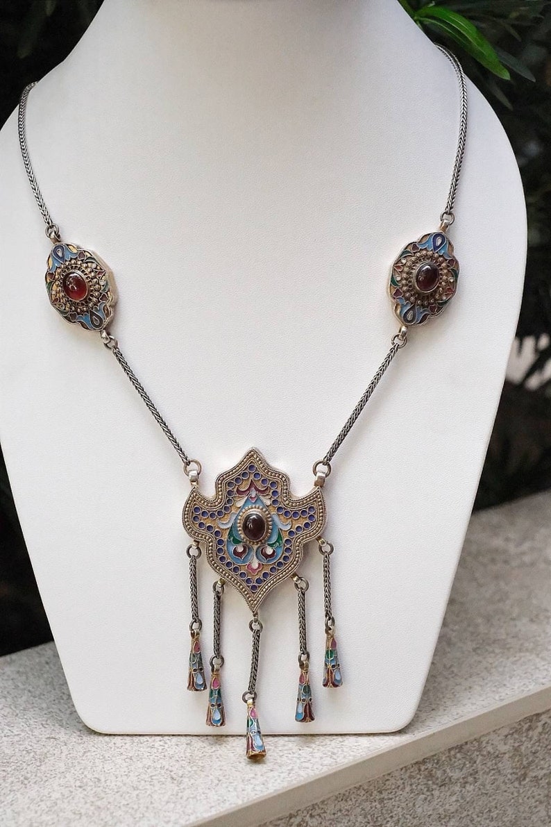 Puede incluir: Un collar de plata con un gran colgante adornado con trabajo de esmalte azul, verde y dorado. El colgante tiene una piedra preciosa roja en el centro y cuelga de una cadena con dos colgantes m&aacute;s peque&ntilde;os a juego.