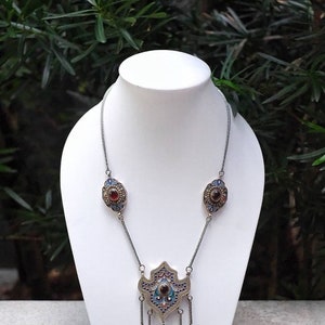 Puede incluir: Un collar de plata con un gran colgante adornado con trabajo de esmalte azul y verde y una piedra preciosa roja. El colgante tiene forma de l&aacute;grima con cadenas colgantes y piedras preciosas m&aacute;s peque&ntilde;as.