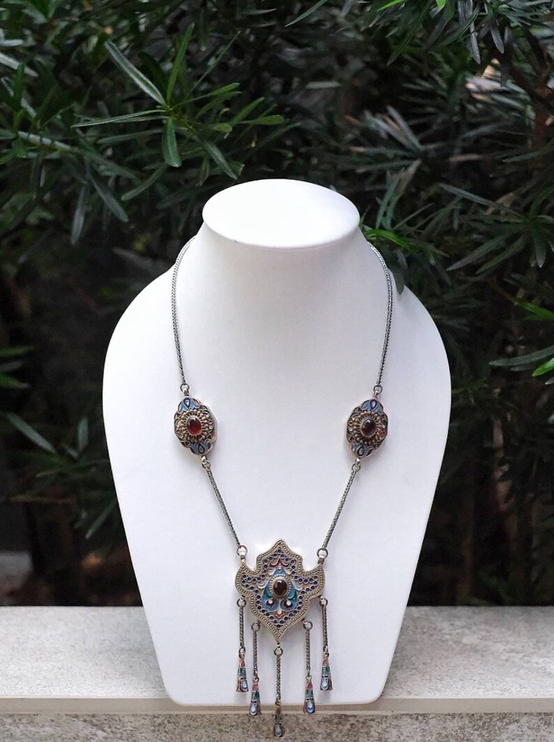 Puede incluir: Un collar de plata con un gran colgante adornado con trabajo de esmalte azul y verde y una piedra preciosa roja. El colgante tiene forma de l&aacute;grima con cadenas colgantes y piedras preciosas m&aacute;s peque&ntilde;as.