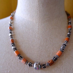 Puede incluir: Un collar con cuentas cilíndricas de color naranja e intercaladas con cuentas y adornos metálicos plateados. Una gran cuenta esférica plateada está centrada en el collar. El collar se exhibe en un maniquí. Las cuentas miden aproximadamente 0,5 cm de diámetro.