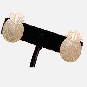 May include: A pair of pineapple-shaped earrings. The earrings are carved with a grid pattern and have a pearlescent finish. The top of the pineapple has a textured, leafy design. The earrings are displayed on a black stand.
